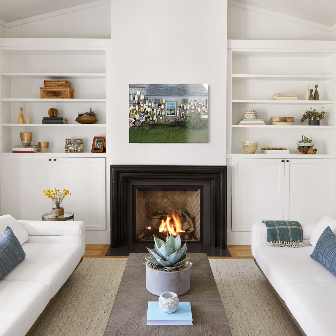 Modern living room with white walls, a fireplace, and a large glass print above it, featuring a coastal house with an assortment of buoys attached to its exterior.