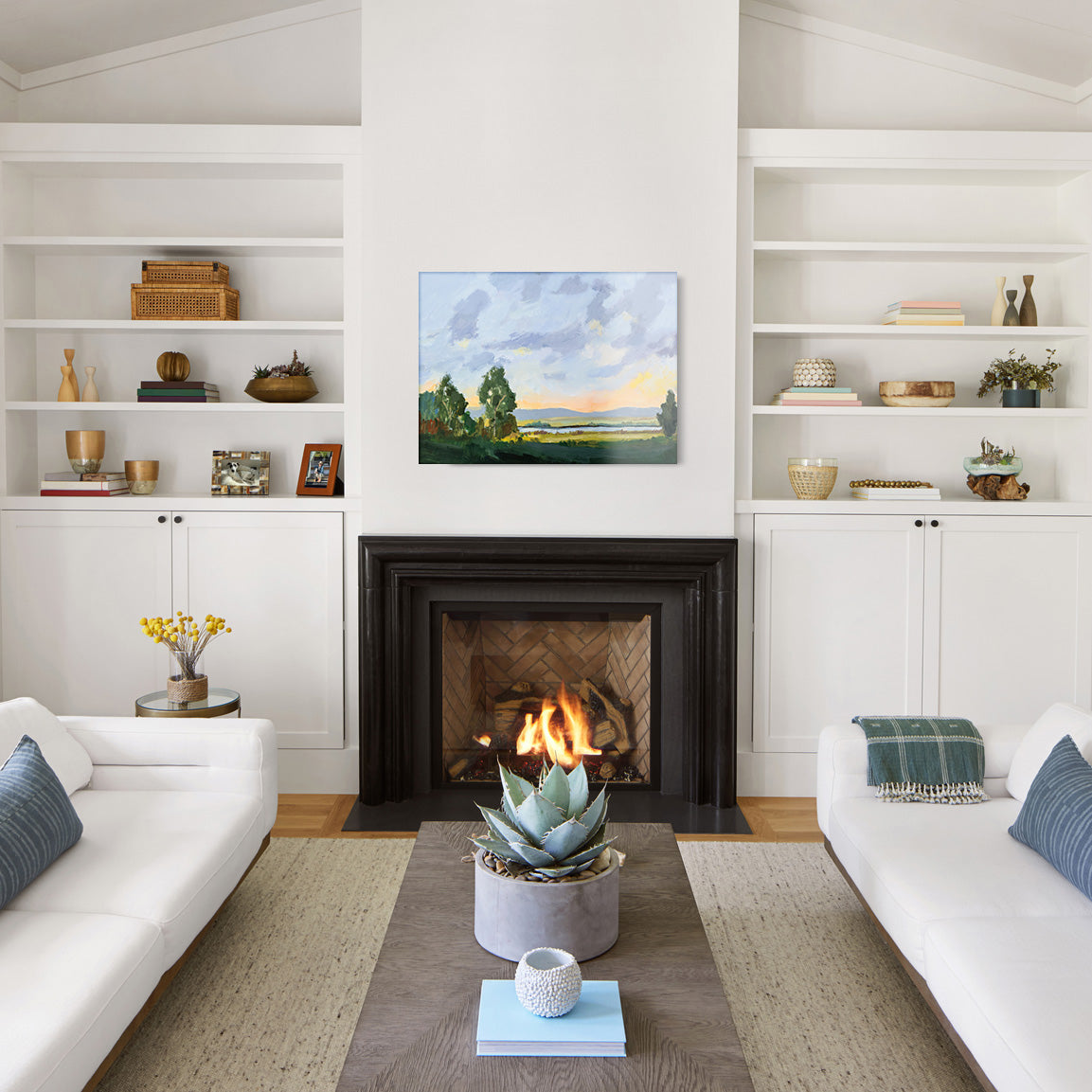 Living room with white walls, a fireplace, and a glass print of a landscape painting with green trees and body of water in the background.
