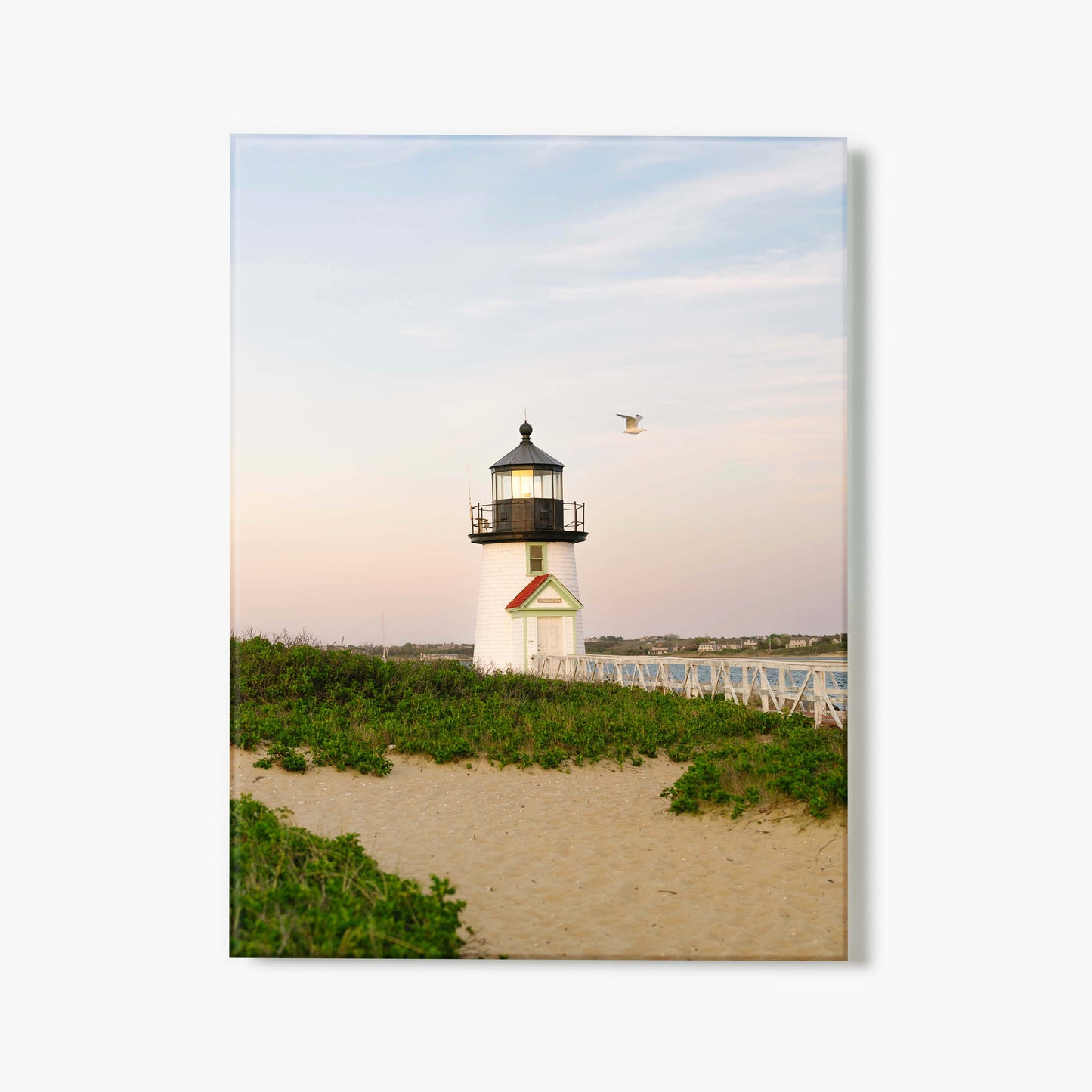 A glass print of a lighthouse on a sandy beach with greenery and a clear sky
