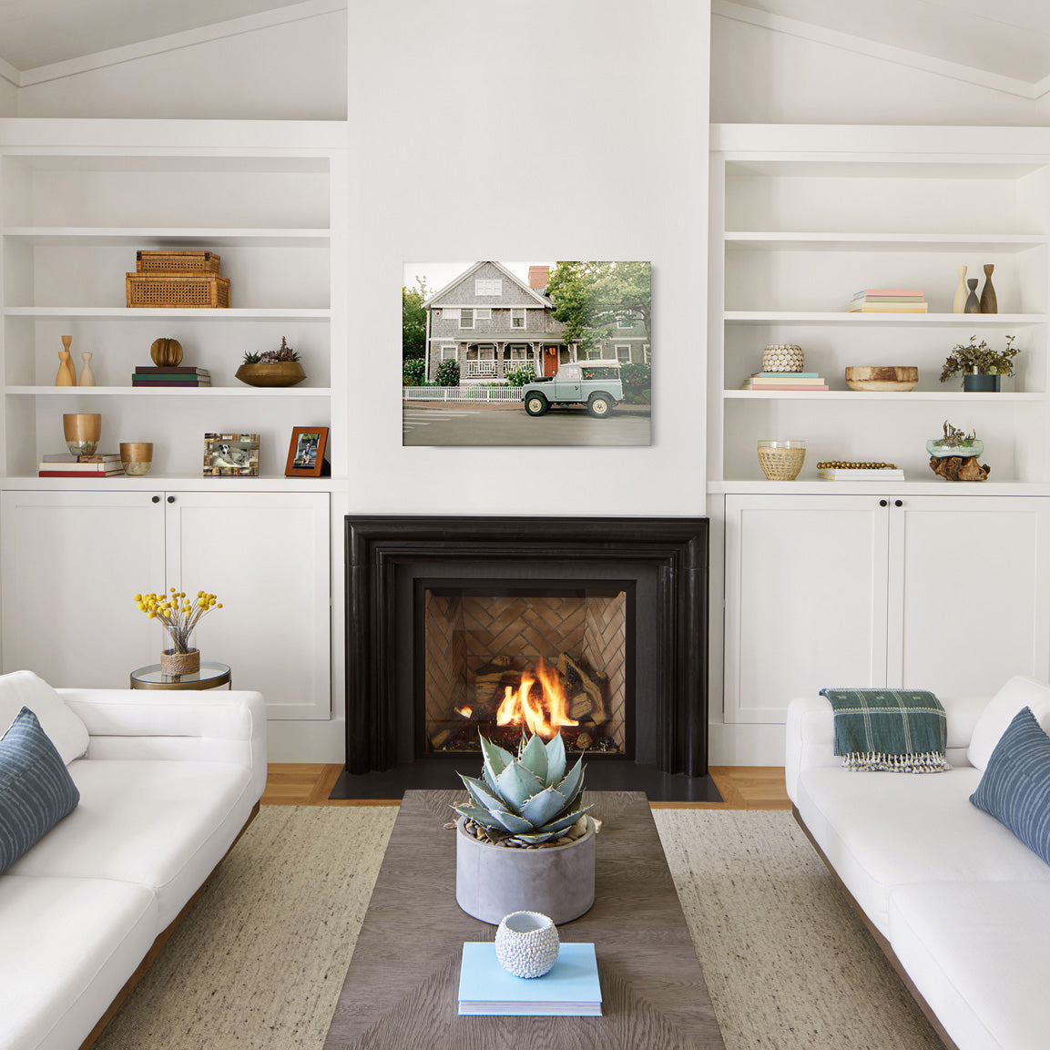 Living room with white walls, a fireplace, and a glass print of a vintage land rover parked in front of a house with white picket fence and flowers.