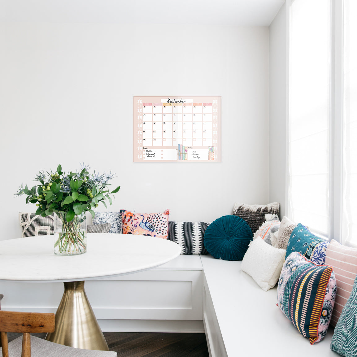 Kitchen nook with a white wall, colorful pillows on a bench, and a pink calendar printed on glass.