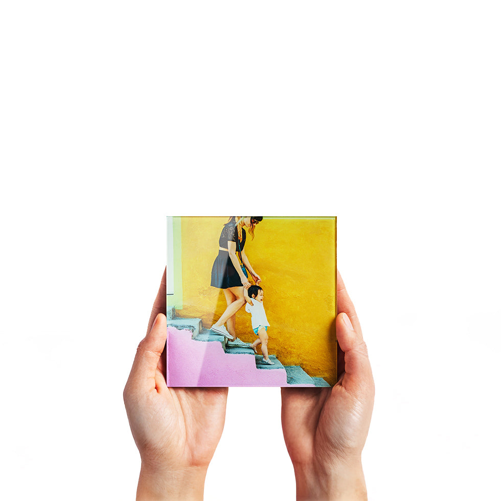 A square glass print being held by hands, featuring a woman and child walking down a pink staircase in front of a yellow wall.