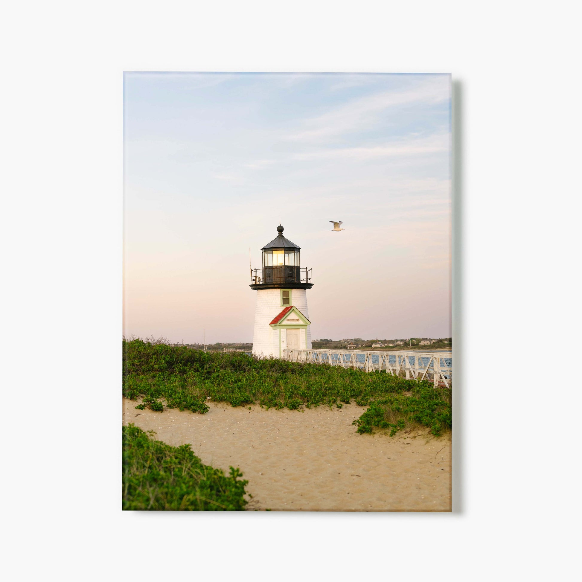 A glass print of a lighthouse on a sandy beach with greenery and a clear sky