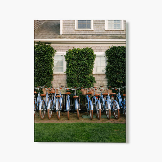 Glass print of a row of bicycles with wooden baskets in front of a house with greenery.