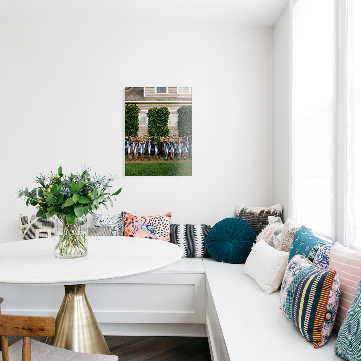 Kitchen with a white bench seating, colorful pillows, a small table with flowers, and a glass print of vintage bikes against an ivy building on the wall