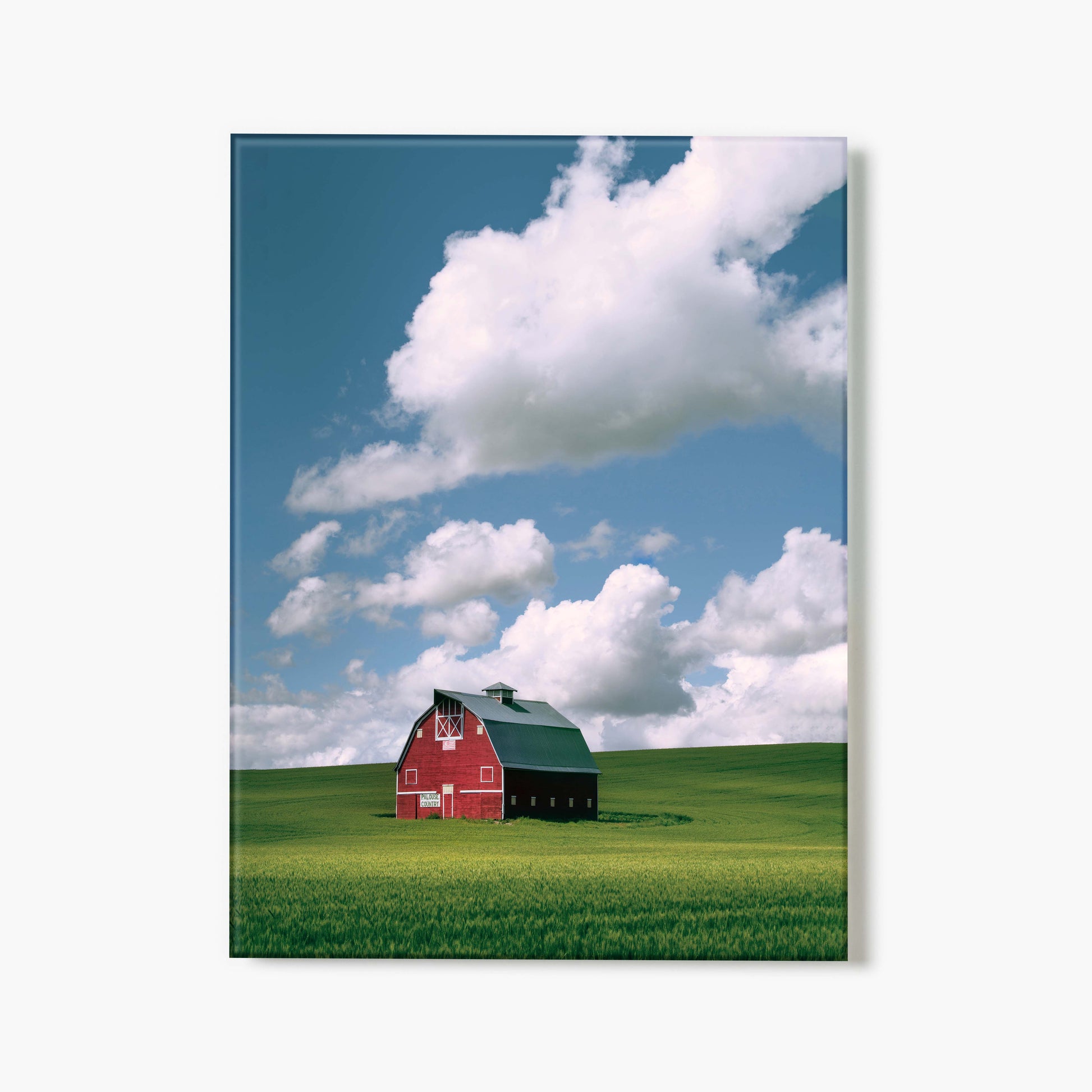 A glass print of a red barn on a green field with a blue sky and white clouds