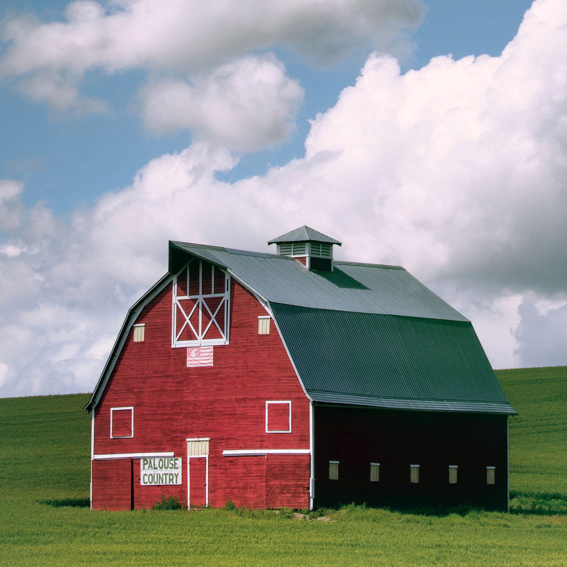 Close up of red barn with green roof on a grassy field under a blue sky with clouds