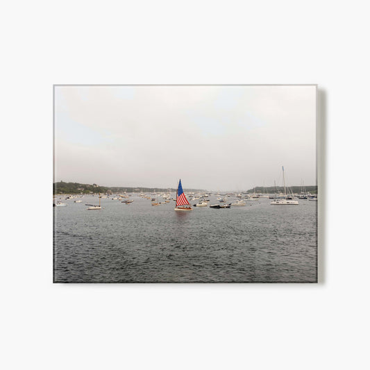 Glass print of a sailboat with an American flag on a body of water with other boats in the background.